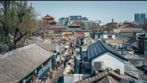 A bustling Beijing Hutong street seen from above, showing traditional tiled rooftops contrasted with modern high-rises in the background