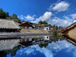 Water reflection of traditional Chinese tiled-roof buildings in the ancient town of Lijiang, Yunnan, China, under a bright blue sky with white clouds.
