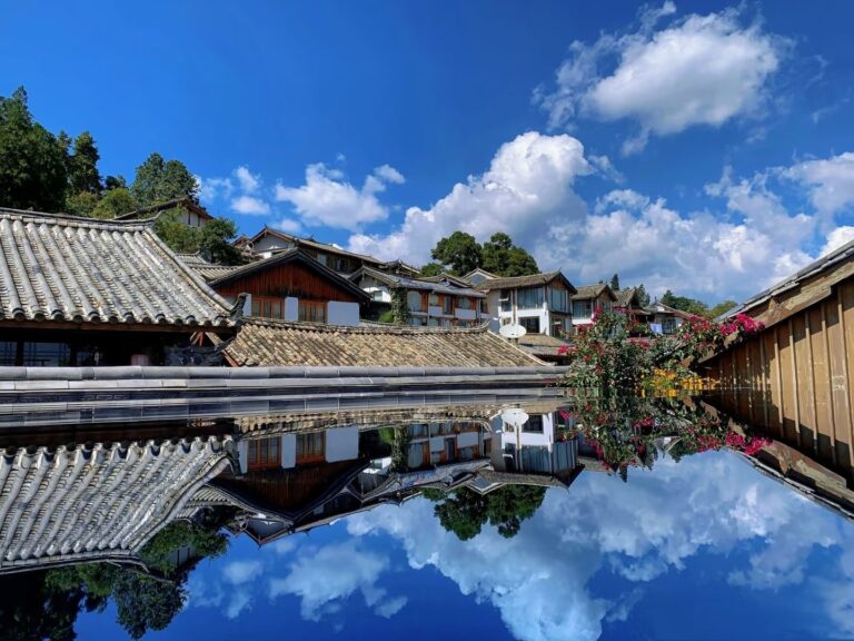 Water reflection of traditional Chinese tiled-roof buildings in the ancient town of Lijiang, Yunnan, China, under a bright blue sky with white clouds.